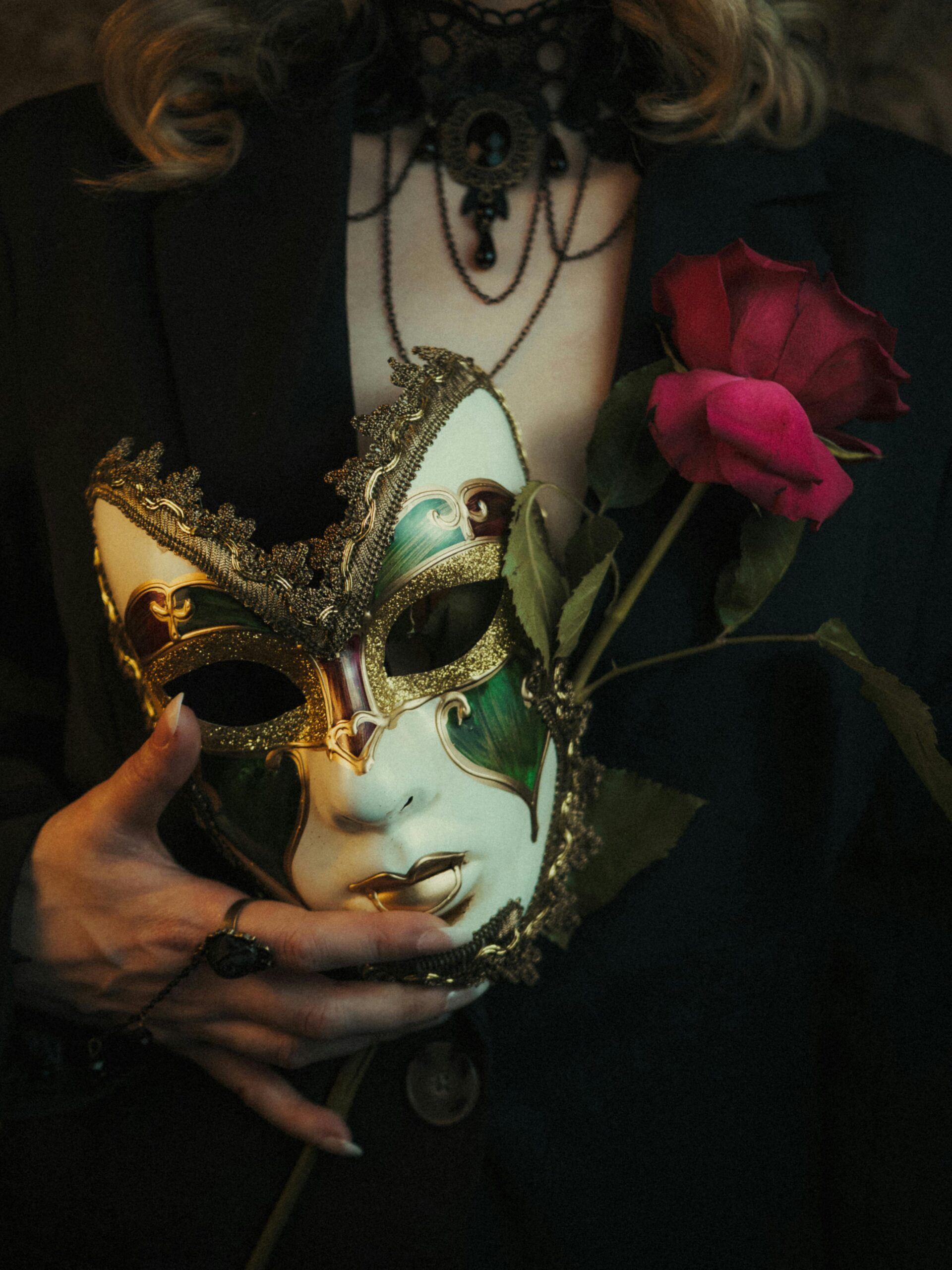 Elegant hand holding a Venetian mask and red rose, capturing mystery and romance in a moody setting.